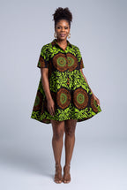 A woman modeling a short-sleeve mini dress with a vibrant green, black, and brown African wax print featuring circular mandala patterns, set against a solid light gray studio background.