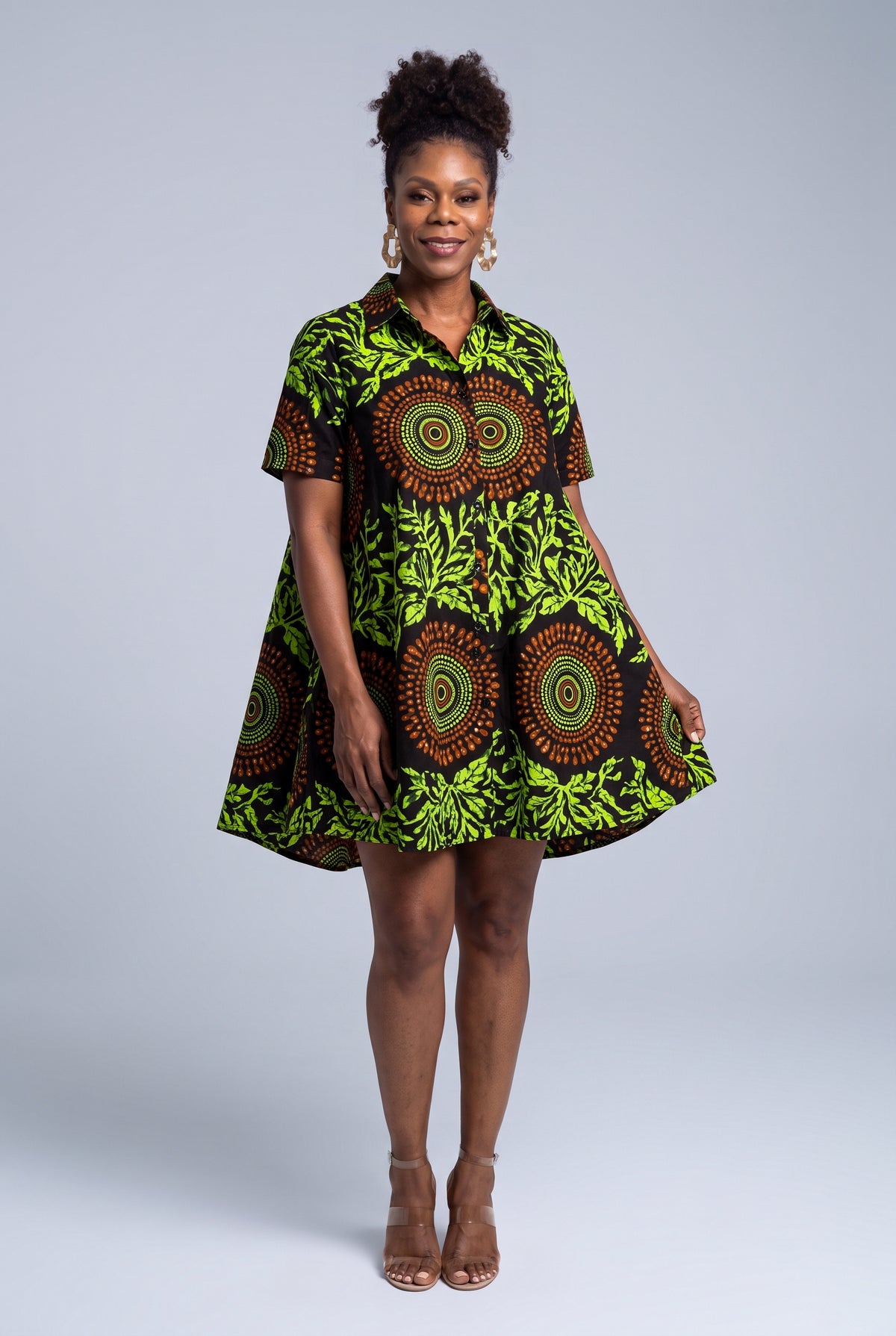 A woman modeling a short-sleeve mini dress with a vibrant green, black, and brown African wax print featuring circular mandala patterns, set against a solid light gray studio background.