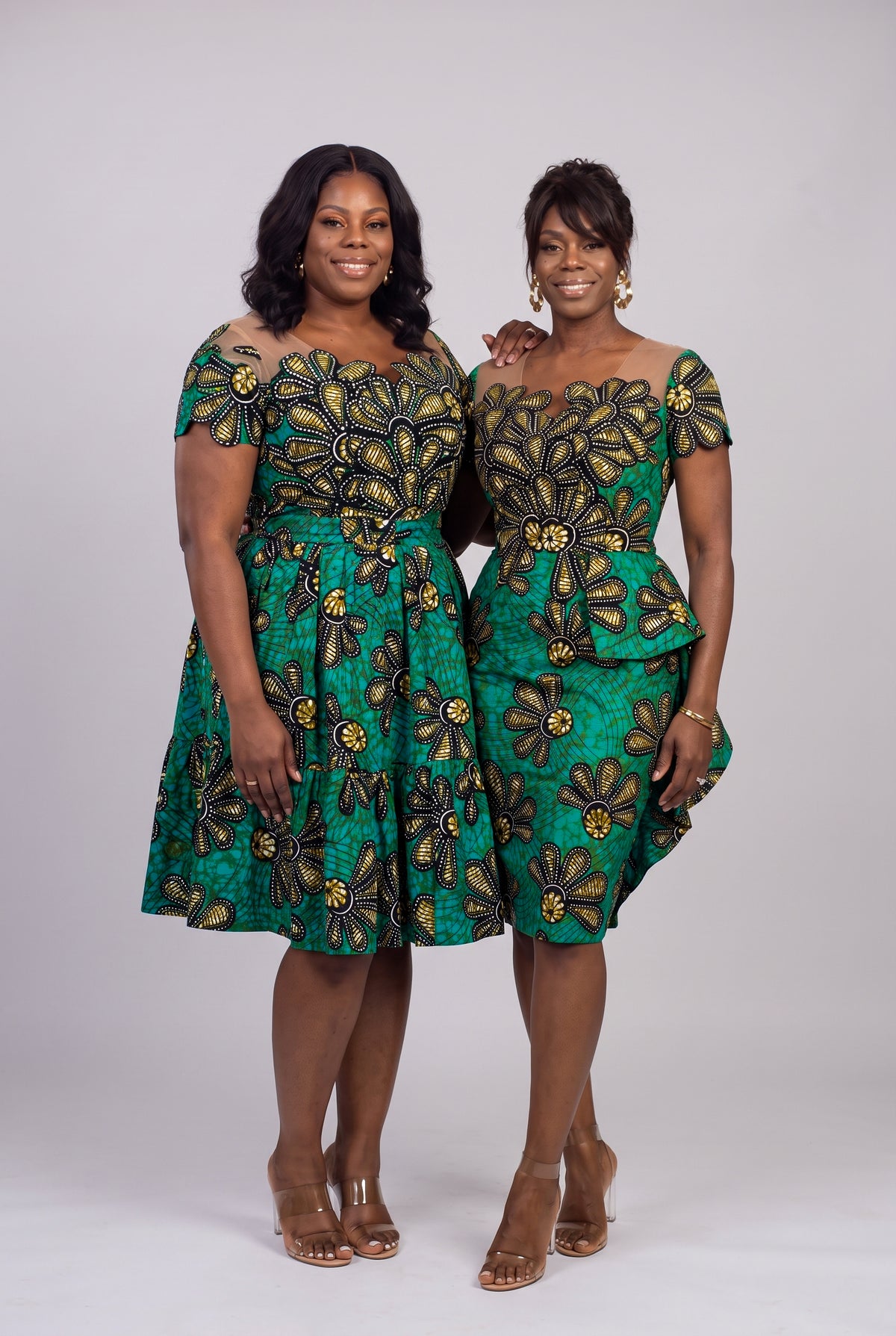 Two women modeling matching green African wax print dresses with black and gold floral patterns