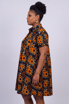 A woman shown in side profile wearing a short-sleeve button-up African shirt dress with an orange and yellow floral print on a black background,