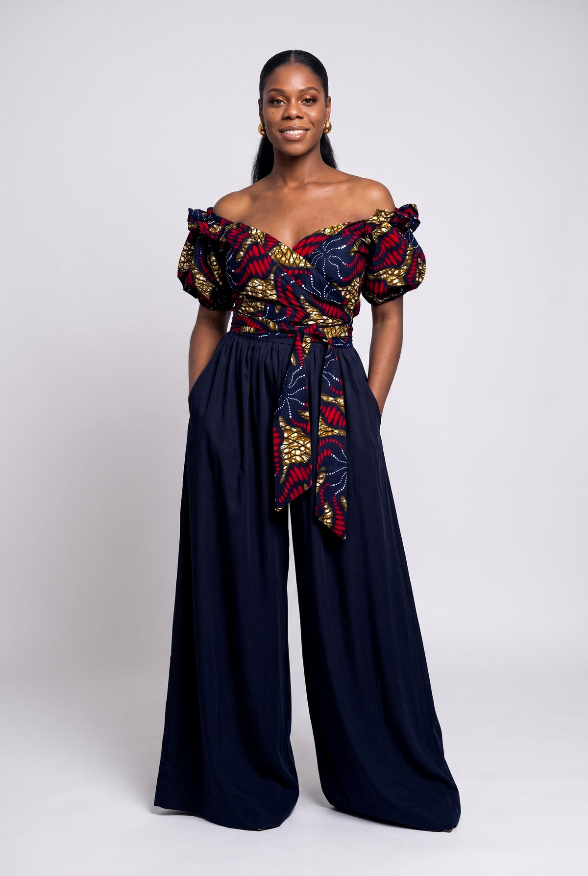 Woman wearing an off-the-shoulder Ankara wrap top in red, gold and black with solid navy blue trousers