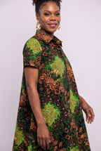 A woman wearing a short-sleeve collared dress with a vibrant green, brown, and black African wax print pattern, photographed against a plain light gray studio background.