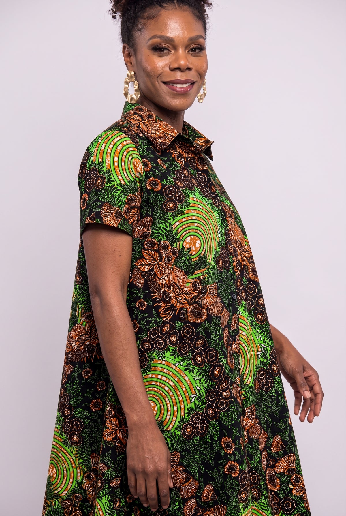 A woman wearing a short-sleeve collared dress with a vibrant green, brown, and black African wax print pattern, photographed against a plain light gray studio background.